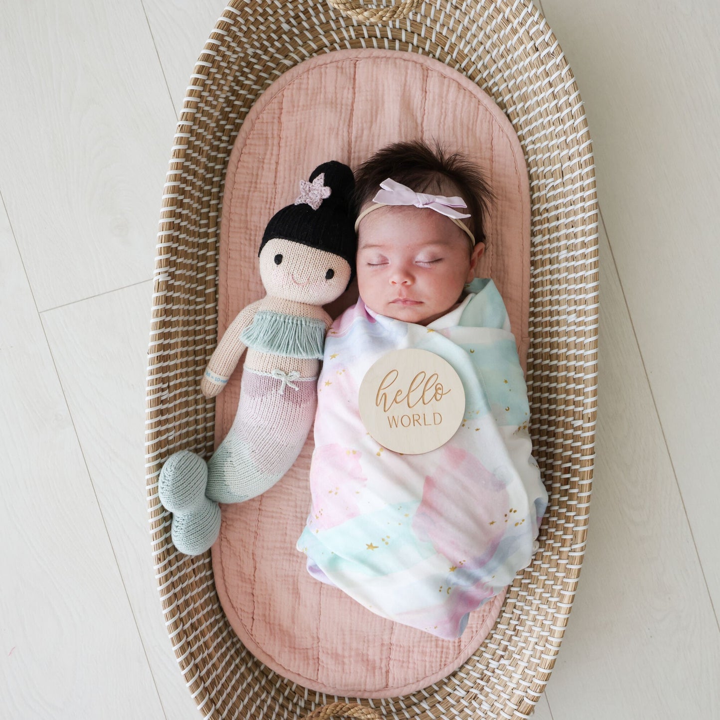 A sleeping baby in a bassinet, snuggled beside Luna the mermaid. There is a small, wooden sign resting on the baby with text that says, “hello world.”