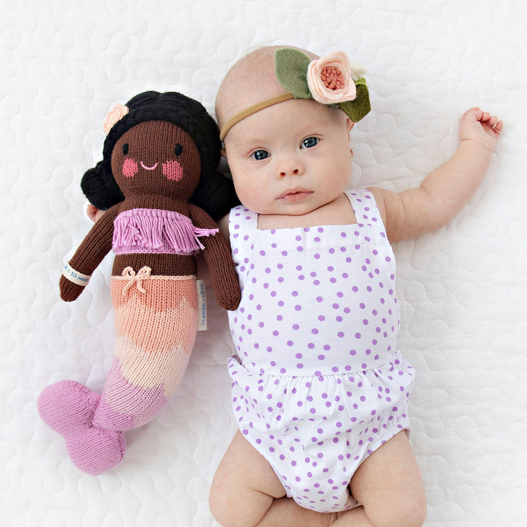 A baby with her arm around a Maya the mermaid doll in the little size.